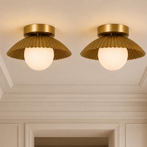 May include: Two flush mount ceiling lights featuring a circular brass canopy and a wide, pleated, metallic gold shade. Each fixture includes a central, spherical, frosted white glass globe that emits a soft, warm glow from the centre.