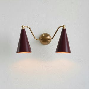 Op de afbeelding: Een wandlamp met twee bordeauxrode, kegelvormige kappen. De armatuur heeft een messing afwerking, met gebogen armen die uit een centrale bol komen. Het licht is aan en geeft een warme gloed.