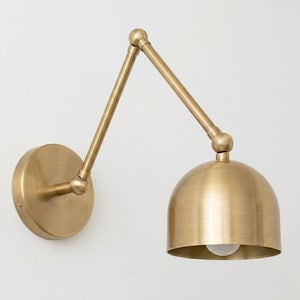 May include: A brass-colored wall-mounted lamp with an adjustable arm and a dome-shaped shade. The lamp is attached to a circular base and has a simple, elegant design. The light fixture is made of metal and has a warm, inviting appearance.