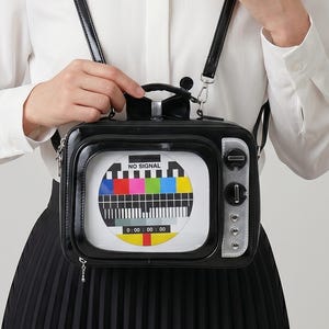 以下が含まれることがあります： 黒色のヴィンテージテレビ型のハンドバッグ。バッグには「NO SIGNAL」の文字とカラフルなテストパターンが表示された画面があります。バッグにはハンドルと調節可能なストラップが付いています。