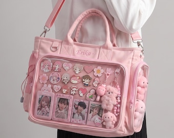 May include: A light pink ita bag with a clear front display window, featuring the name Erika embroidered on the top panel. The bag is filled with various character pins, small plush charms, and four rectangular photo cards inside the clear pocket.