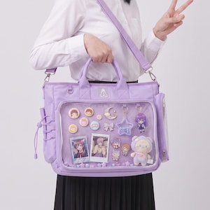 May include: A light purple ita bag featuring a large clear front display window filled with various enamel pins, two small rectangular photo prints, a star-shaped charm, and a small plush character toy. The bag has top handles and a shoulder strap.