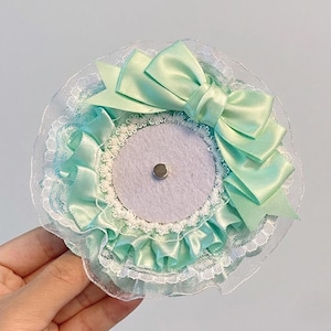 Könnte beinhalten: Ein mintgrünes und weißes dekoratives Haaraccessoire. Das Accessoire verfügt über eine große Satinschleife, Rüschenbesatz und zarte Spitzenkanten. In der Mitte befindet sich ein kleiner, silberfarbener Magnet. Das Accessoire wird von einer Hand gehalten.