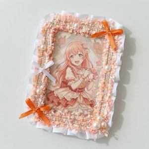 Op de afbeelding: Een rechthoekige fotolijst versierd met perzikkleurige textuurrand, witte ruches, kleine witte parels en twee oranje satijnen strikken. Binnenin zit een anime-stijl illustratie van een glimlachend persoon met lang perzikkleurig haar die een theekopje vasthoudt.