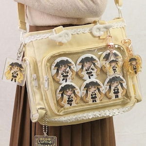 May include: A light yellow shoulder bag with a clear front pocket displaying several round character pins. The bag has a lace trim, cloud-shaped accents, and attached keychains. The bag also has a side pocket with a zipper.