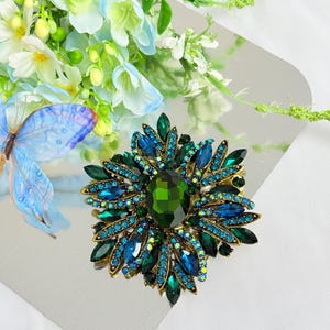May include: A large, ornate brooch with a floral design. The brooch features a central, oval-shaped green gemstone surrounded by blue and green crystals. The setting is gold-toned, and the brooch is set against a silver background with floral accents.