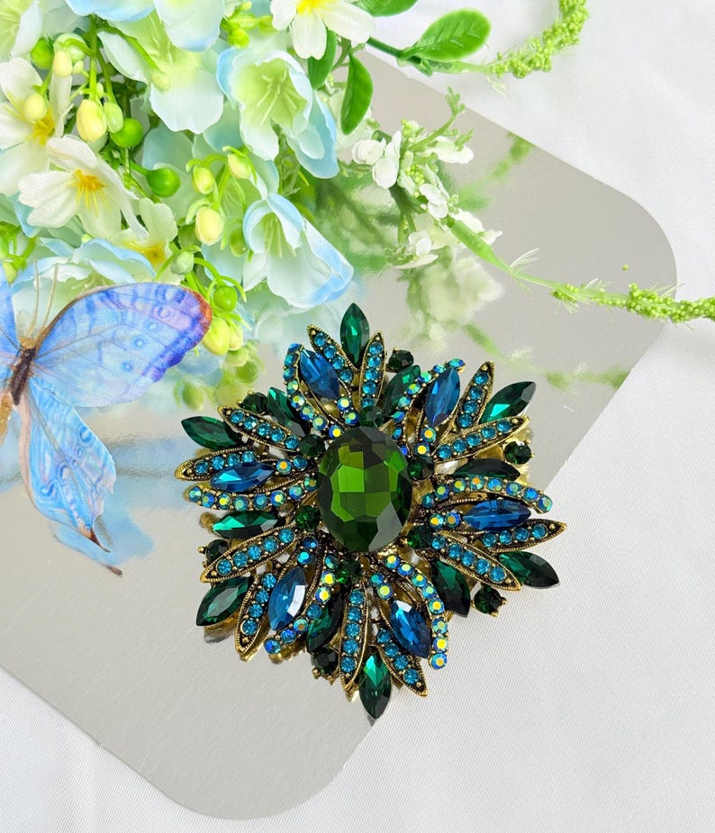 May include: A large, ornate brooch with a floral design. The brooch features a central, oval-shaped green gemstone surrounded by blue and green crystals. The setting is gold-toned, and the brooch is set against a silver background with floral accents.