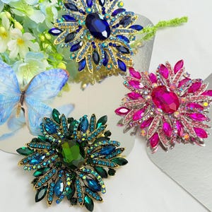May include: Three jeweled brooches in shades of blue, green, and pink. Each brooch features a large central stone surrounded by smaller, colorful gems. The brooches are set on a gold-toned metal base, creating a sparkling, eye-catching design.
