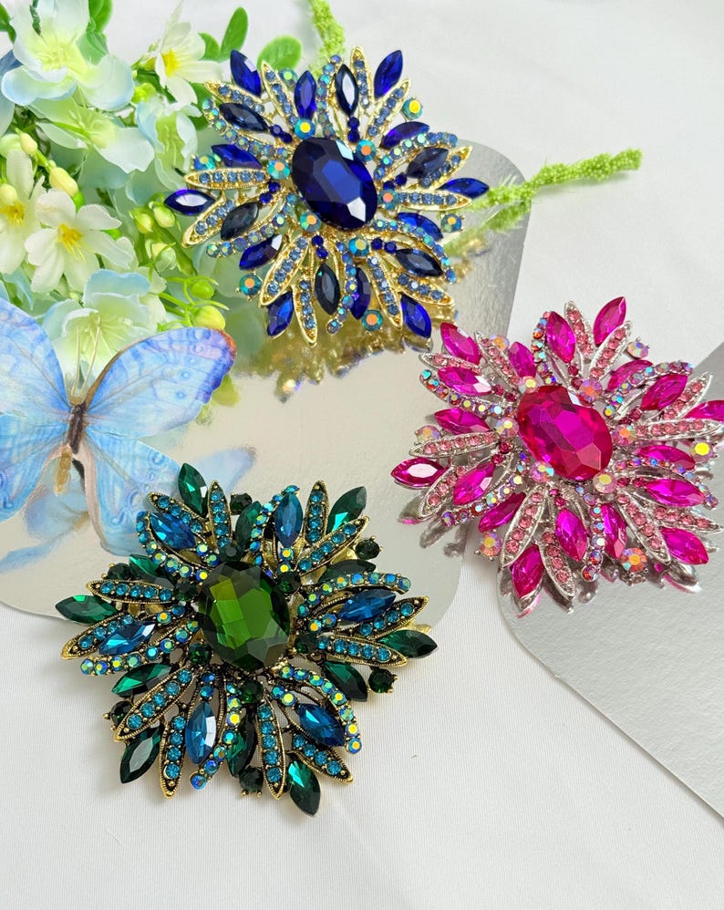 May include: Three jeweled brooches in shades of blue, green, and pink. Each brooch features a large central stone surrounded by smaller, colorful gems. The brooches are set on a gold-toned metal base, creating a sparkling, eye-catching design.