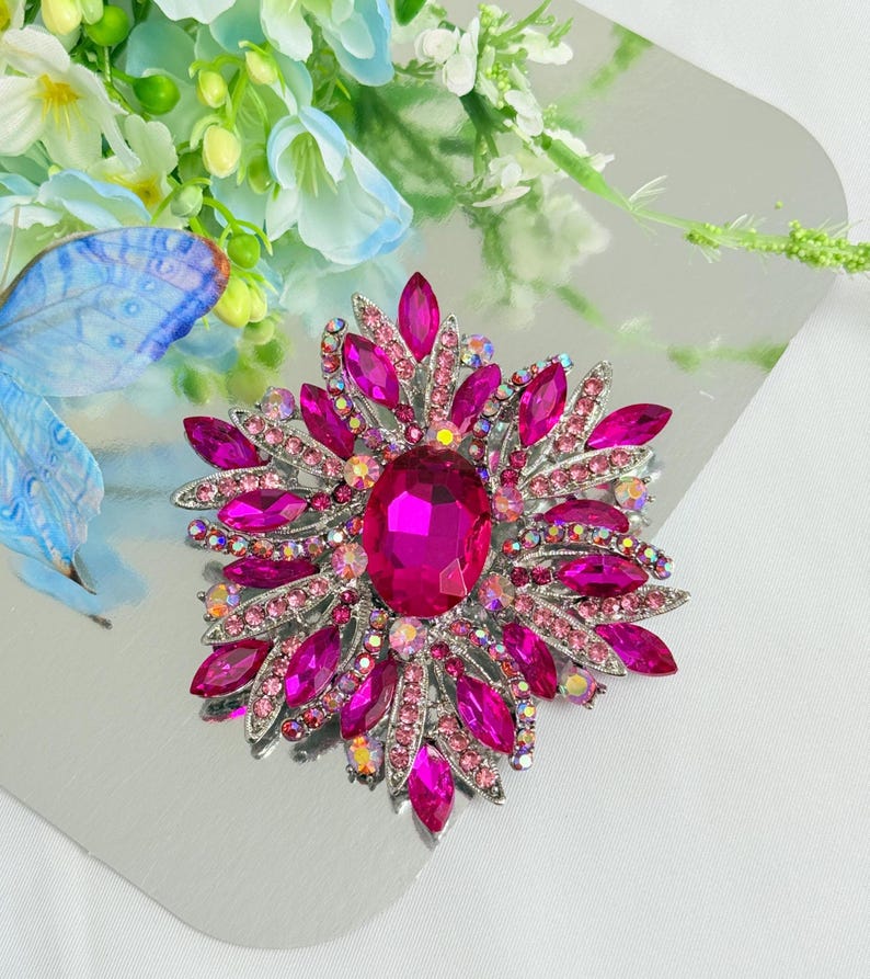 May include: A vibrant, eye-catching brooch featuring a large, oval, fuchsia-colored center stone surrounded by smaller pink and clear crystals. The brooch has a silver-toned metal setting and a floral design, perfect for adding a touch of glamour.