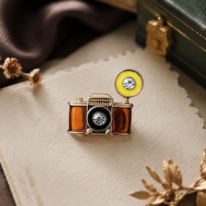 May include: A gold-toned camera brooch featuring brown enamel side panels, a black lens housing with a central clear rhinestone, and a yellow circular flash attachment topped with a matching clear rhinestone accent.