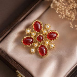 May include: A gold-toned brooch with a floral design. The brooch features large, oval, red gemstones surrounded by smaller clear stones. Small, round, white pearls are interspersed throughout the design. The brooch is displayed on a satin surface.