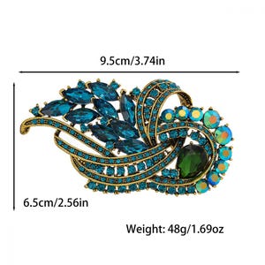 K&ouml;nnte beinhalten: Verzierte Brosche mit goldfarbener Metallbasis, verziert mit t&uuml;rkisblauen und irisierenden Kristallen. Die Brosche ist 9,5 cm lang und 6,5 cm breit und wiegt 48 g. Sie hat einen gro&szlig;en gr&uuml;nen Mittelstein.