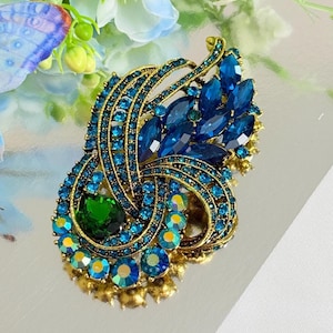 May include: Ornate gold-toned brooch with a peacock feather design. The brooch features a large, oval emerald-green stone at the center, surrounded by intricate detailing and numerous blue and teal crystals. The brooch is a statement piece.