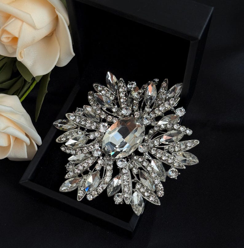 May include: A silver-toned brooch with a large, oval-shaped clear crystal at its center, surrounded by numerous smaller, clear crystals. The brooch is displayed in a black box, with a cream-colored rose in the background.