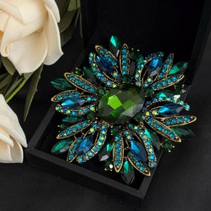 May include: A large, ornate brooch with a floral design. The brooch features a central, faceted, emerald-green stone surrounded by petals of teal and blue crystals. The setting is gold-toned, and the brooch is displayed in a black box.
