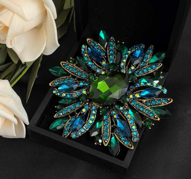 May include: A large, ornate brooch with a floral design. The brooch features a central, faceted, emerald-green stone surrounded by petals of teal and blue crystals. The setting is gold-toned, and the brooch is displayed in a black box.