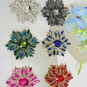 May include: A collection of six jeweled flower brooches in various colors. The brooches feature a central gemstone surrounded by smaller stones, creating a floral design. Colors include silver, black, green, blue, pink, and red.
