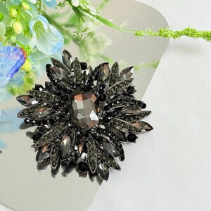 May include: A large, dark gray brooch with a central, faceted, oval-shaped stone. The brooch is surrounded by multiple layers of smaller, dark gray, and black stones, creating a floral design. The brooch is set on a silver surface.