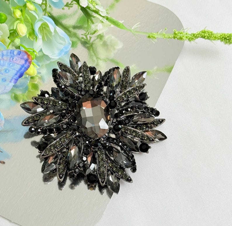 May include: A large, dark gray brooch with a central, faceted, oval-shaped stone. The brooch is surrounded by multiple layers of smaller, dark gray, and black stones, creating a floral design. The brooch is set on a silver surface.