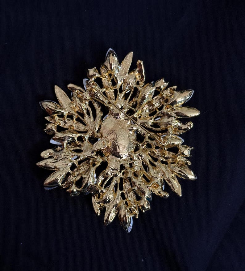 May include: A gold-toned brooch with a floral design. The back of the brooch features a pin for attachment. The brooch has a detailed, textured surface with a mix of shiny and matte finishes. The brooch is set against a dark background.