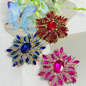 May include: Three jeweled brooches in red, blue, and pink. Each brooch features a large central stone surrounded by smaller gems, creating a floral or starburst design. The brooches are set on a reflective surface.