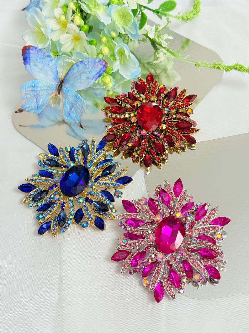 May include: Three jeweled brooches in red, blue, and pink. Each brooch features a large central stone surrounded by smaller gems, creating a floral or starburst design. The brooches are set on a reflective surface.