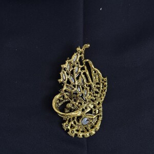 K&ouml;nnte beinhalten: Eine goldfarbene Brosche mit einem aufwendigen, durchbrochenen Design. Die Brosche hat eine zentrale Ansammlung klarer, funkelnder Steine und ist vor einem dunklen Hintergrund platziert. Der Anstecker ist ein dekoratives Accessoire.