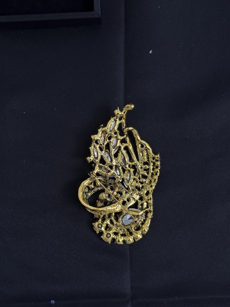K&ouml;nnte beinhalten: Eine goldfarbene Brosche mit einem aufwendigen, durchbrochenen Design. Die Brosche hat eine zentrale Ansammlung klarer, funkelnder Steine und ist vor einem dunklen Hintergrund platziert. Der Anstecker ist ein dekoratives Accessoire.