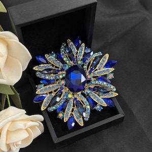 May include: A dazzling blue and gold brooch, shaped like a flower, is displayed in a black box. The brooch features a large, oval, dark blue center stone surrounded by smaller blue and iridescent crystals, set in a gold-toned metal frame.