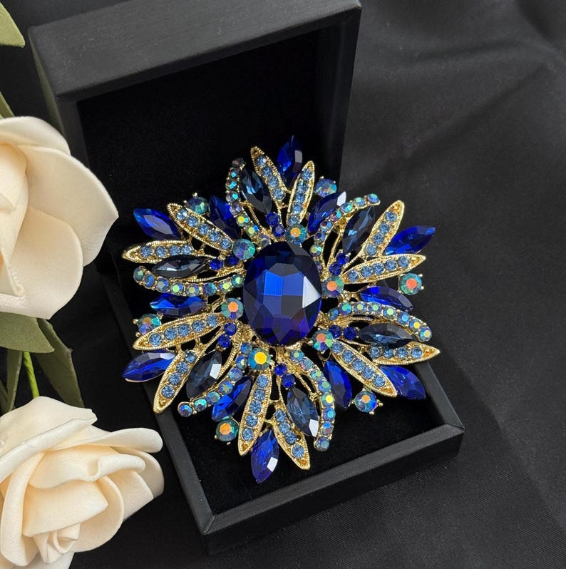 May include: A dazzling blue and gold brooch, shaped like a flower, is displayed in a black box. The brooch features a large, oval, dark blue center stone surrounded by smaller blue and iridescent crystals, set in a gold-toned metal frame.