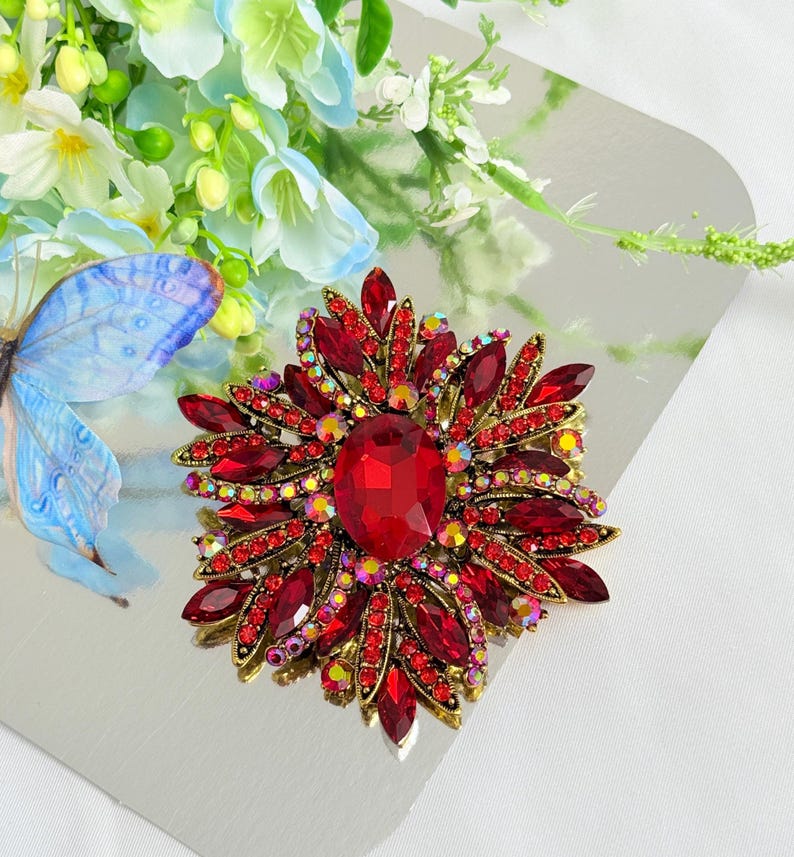 May include: A large, ornate brooch with a floral design. The brooch features a central, oval-shaped red gemstone surrounded by numerous red and iridescent crystals. The setting is gold-toned.