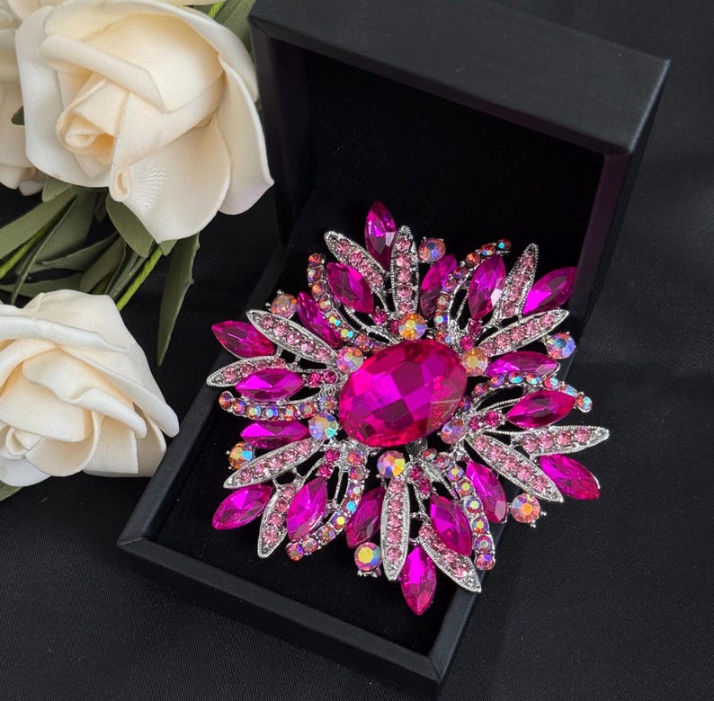 May include: A large, ornate brooch with a floral design. The brooch features a large, oval, magenta-colored center stone surrounded by smaller, pink and magenta crystals. The brooch is set in a silver-toned metal and is displayed in a black box.
