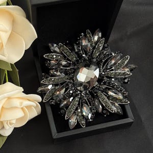 May include: A large, ornate brooch with a floral design. The brooch features a central, oval-shaped, faceted gray crystal surrounded by numerous smaller, dark gray crystals. It is set in a black metal frame and presented in a black box.