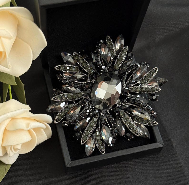 May include: A large, ornate brooch with a floral design. The brooch features a central, oval-shaped, faceted gray crystal surrounded by numerous smaller, dark gray crystals. It is set in a black metal frame and presented in a black box.