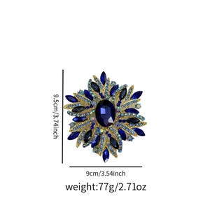 May include: A gold-toned brooch with a large, oval, dark blue gemstone at its center, surrounded by smaller blue and clear crystals. The brooch measures 3.74 inches in height and 3.54 inches in width, and weighs 2.71 ounces.