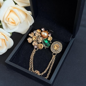 May include: A decorative brooch with a cluster of gemstones in shades of gold, green, and peach, set in a gold-tone metal. The brooch is presented in a black box, with a gold chain and an additional oval-shaped gemstone element.