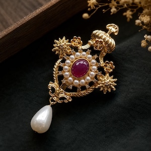 May include: A gold-tone brooch featuring a central oval-shaped deep pink stone surrounded by a ring of small white pearls. The metal frame includes star and flower accents, a crown-like top, and a single dangling white teardrop-shaped pearl.