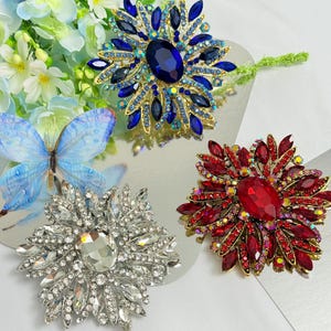 May include: Three jeweled flower brooches in shades of blue, red, and clear crystals. The brooches have a central stone surrounded by smaller gems, creating a floral design. The brooches are set on a reflective surface.