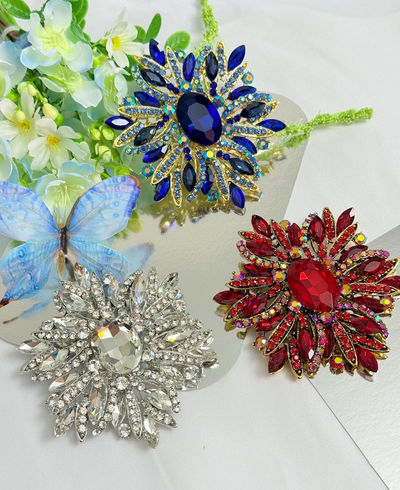 May include: Three jeweled flower brooches in shades of blue, red, and clear crystals. The brooches have a central stone surrounded by smaller gems, creating a floral design. The brooches are set on a reflective surface.