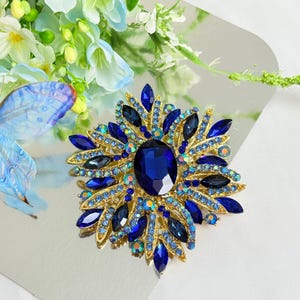 May include: A gold-toned brooch with a large, oval, deep blue center stone, surrounded by smaller blue and iridescent crystals. The brooch has a floral or starburst design, with pointed blue crystal accents radiating outwards.