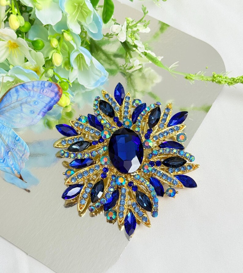 May include: A gold-toned brooch with a large, oval, deep blue center stone, surrounded by smaller blue and iridescent crystals. The brooch has a floral or starburst design, with pointed blue crystal accents radiating outwards.