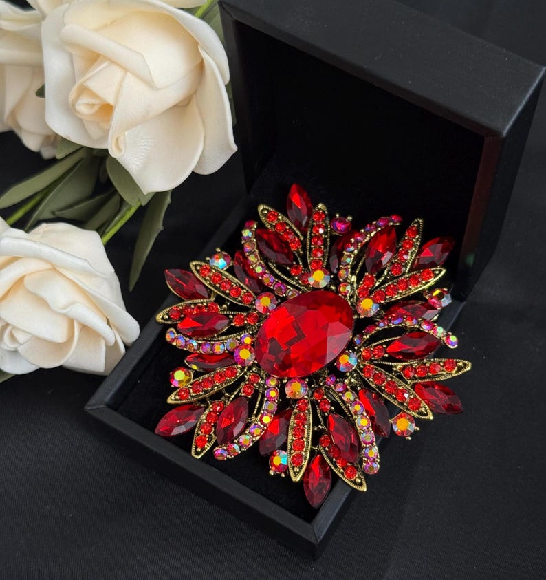 May include: A large, ornate red brooch with a central oval-shaped stone and surrounding smaller red and iridescent crystals. The brooch is set in a gold-toned metal and displayed in a black box, with white roses in the background.