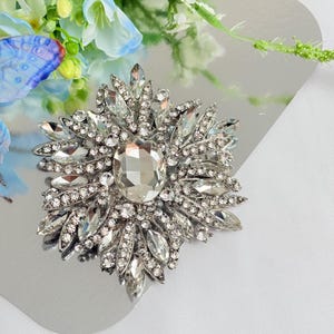 May include: A silver-toned brooch with a central, large, clear, oval-cut crystal surrounded by smaller, clear, marquise-cut crystals. The brooch has a starburst design and is set on a reflective surface.