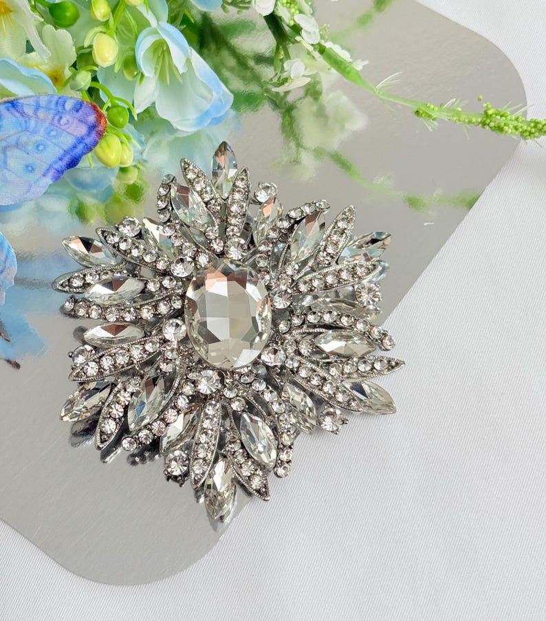 May include: A silver-toned brooch with a central, large, clear, oval-cut crystal surrounded by smaller, clear, marquise-cut crystals. The brooch has a starburst design and is set on a reflective surface.