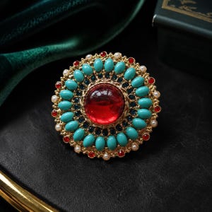 May include: A large circular statement ring featuring a prominent central red cabochon stone surrounded by a halo of oval turquoise-colored beads, small dark blue crystals, tiny white pearls, and small red crystals set in a gold-tone metal base.