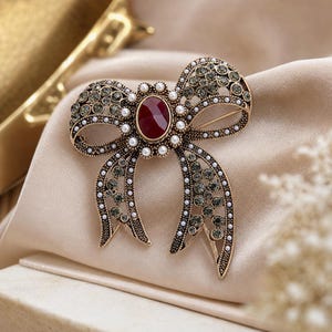 Peut inclure: Une broche en forme de nœud de couleur or vieilli, ornée d'une pierre ovale rouge foncé au centre entourée de petites perles blanches. La surface est agrémentée de nombreux cristaux gris ronds sertis dans la structure métallique du nœud.