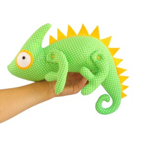 May include: A green polka-dot fabric chameleon plush toy with yellow accents. The stuffed animal has a curved tail, a spiky yellow dorsal fin, and a large eye with an orange border. The toy is held in a hand.
