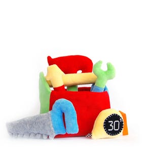 Puede incluir: Una caja de herramientas de juguete con varias herramientas. La caja de herramientas roja contiene un martillo amarillo, una llave inglesa verde y una sierra gris con un mango azul. También se incluye una cinta métrica amarilla con el número 30.