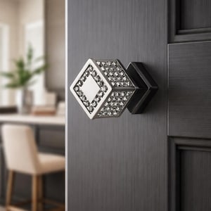 May include: A decorative door knob with a square, diamond-shaped design. The knob has a silver frame with a central square and is encrusted with small crystals. It is mounted on a dark grey door.